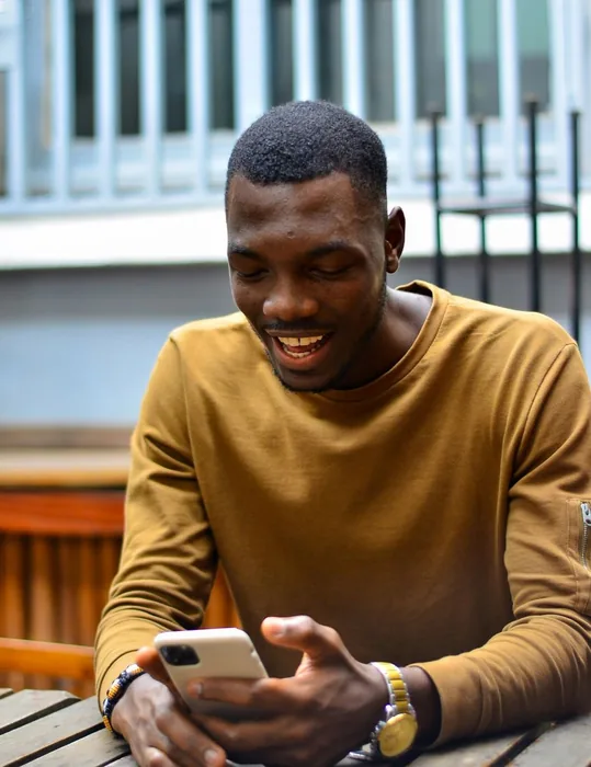 A young man laughing at his phone at a cafe table.