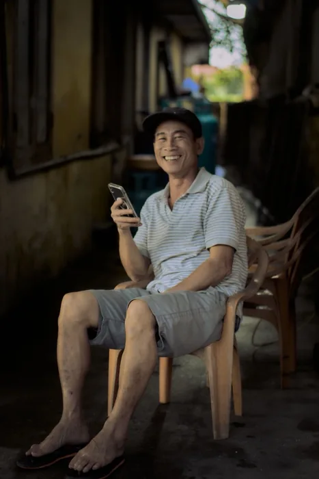 An older man laughing while looking at his phone, seated outside.