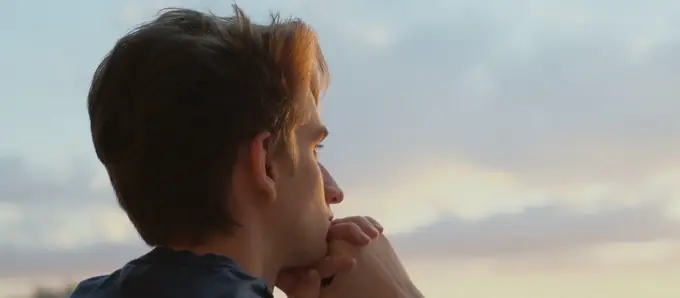 A man at a balcony rail at dusk, chin resting on his hands, looking out.