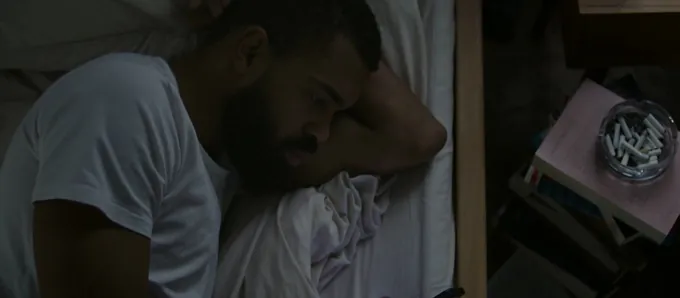 A man sitting on the edge of a bed at night, looking down at the phone in his hands, his reflection in a dark window doing the same.