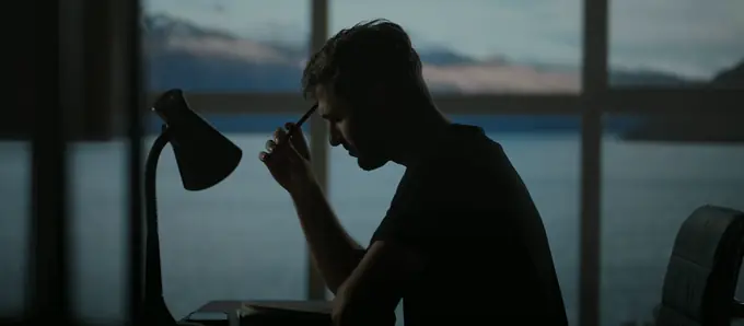A man in silhouette at a desk beside a lamp, a pen at his temple, with dusk over water in the window behind him.