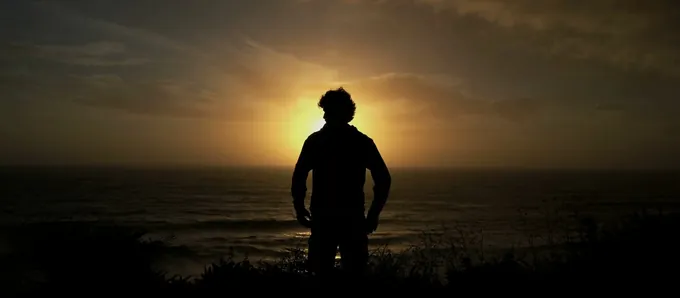 A man standing still at the shore, facing into a low sun.