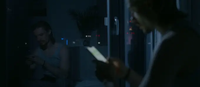 A man sitting on the edge of a bed at night, looking down at the phone in his hands, his reflection in a dark window doing the same.